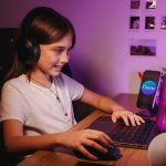 A smiling young girl wearing headphones using a laptop for creative activities, indoors.